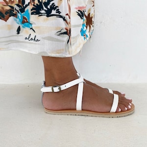 May include: A white flat sandal with thin straps, an ankle strap featuring a metallic buckle, and a tan sole. The sandal is worn with a white fabric garment featuring a tropical print and the word aloha written in black cursive script.