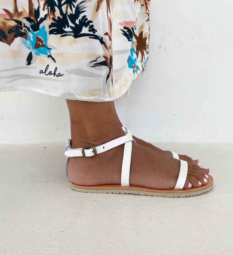 May include: A white flat sandal with thin straps, an ankle strap featuring a metallic buckle, and a tan sole. The sandal is worn with a white fabric garment featuring a tropical print and the word aloha written in black cursive script.