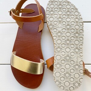 Peut inclure: Une paire de sandales avec une semelle int&eacute;rieure en cuir marron, une bride avant en cuir dor&eacute; m&eacute;tallis&eacute; et une bride de cheville en cuir marron avec une boucle en laiton. La semelle ext&eacute;rieure en caoutchouc gris clair est textur&eacute;e.