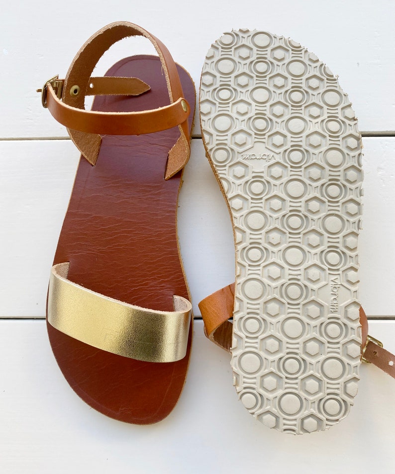 Peut inclure: Une paire de sandales avec une semelle int&eacute;rieure en cuir marron, une bride avant en cuir dor&eacute; m&eacute;tallis&eacute; et une bride de cheville en cuir marron avec une boucle en laiton. La semelle ext&eacute;rieure en caoutchouc gris clair est textur&eacute;e.