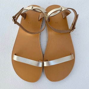May include: A pair of flat sandals featuring metallic gold straps across the toes and around the ankles with adjustable buckles. The footbeds are tan with the number 36 printed in the center. The straps are secured with gold-toned metal hardware.