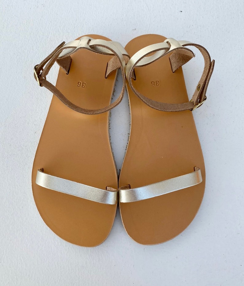 May include: A pair of flat sandals featuring metallic gold straps across the toes and around the ankles with adjustable buckles. The footbeds are tan with the number 36 printed in the center. The straps are secured with gold-toned metal hardware.