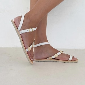 May include: White flat sandals featuring thin straps, a gold-tone buckle closure at the ankle, and small circular gold-tone decorative accents along the top strap. The sandals have a flat sole and a toe loop design for a secure fit.