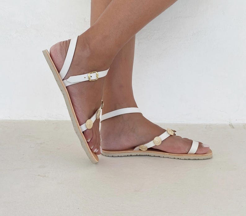 May include: White flat sandals featuring thin straps, a gold-tone buckle closure at the ankle, and small circular gold-tone decorative accents along the top strap. The sandals have a flat sole and a toe loop design for a secure fit.
