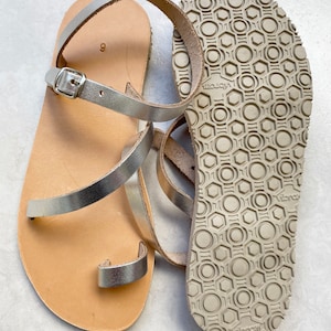 May include: A pair of silver metallic sandals with a toe loop and an adjustable ankle strap with a silver buckle. The footbed is tan, and the outsole features a beige rubber Vibram tread pattern with a repeating hexagonal and circular design.