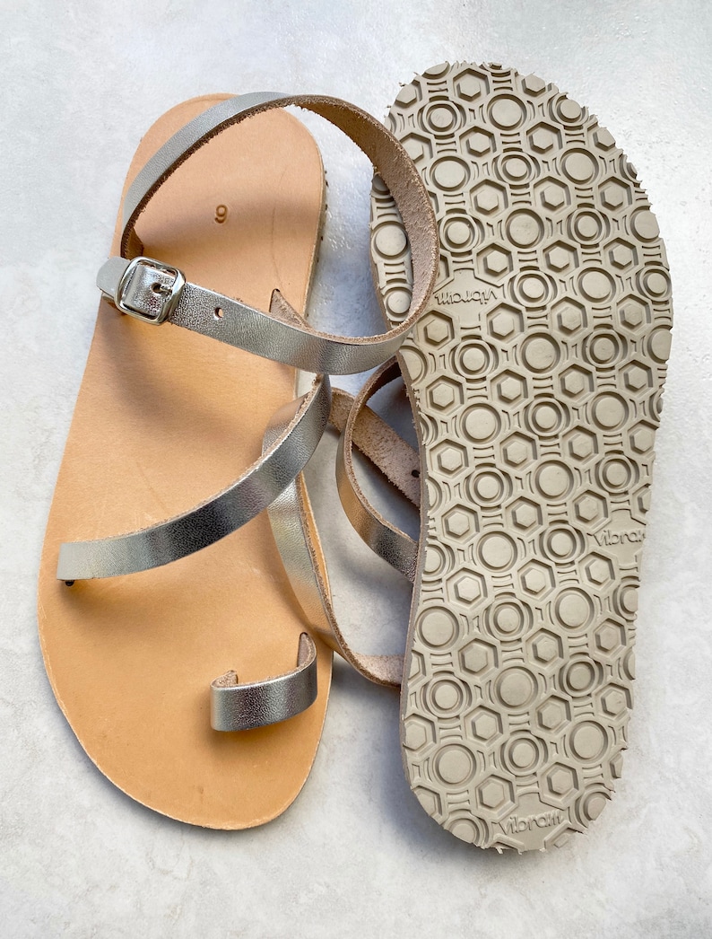 May include: A pair of silver metallic sandals with a toe loop and an adjustable ankle strap with a silver buckle. The footbed is tan, and the outsole features a beige rubber Vibram tread pattern with a repeating hexagonal and circular design.