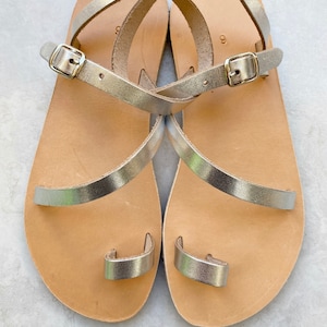 May include: A pair of flat sandals featuring metallic gold straps, a toe loop, and an adjustable ankle strap with a gold-tone metal buckle. The footbed is made of light tan leather, and the number 9 is clearly visible on the heel area of each sandal.