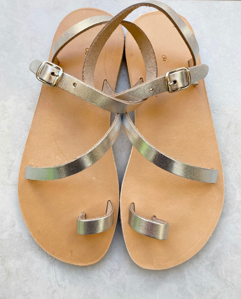 May include: A pair of flat sandals featuring metallic gold straps, a toe loop, and an adjustable ankle strap with a gold-tone metal buckle. The footbed is made of light tan leather, and the number 9 is clearly visible on the heel area of each sandal.