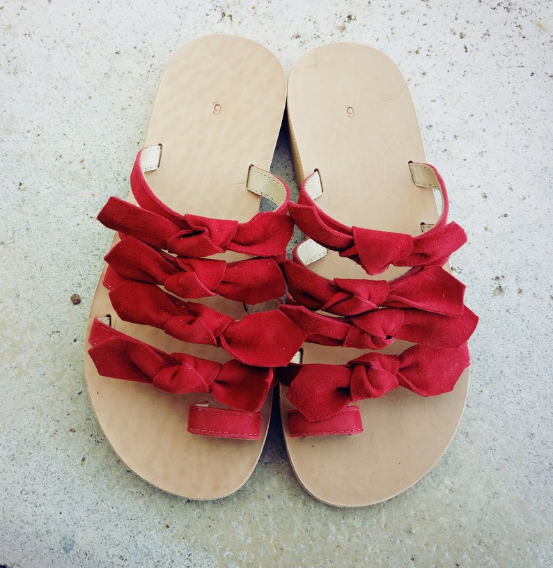 May include: A pair of sandals with red bows. The sandals are made of leather and have a flat sole.