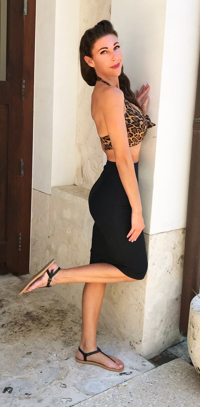 A woman wearing a black skirt and a leopard print halter top. She is standing with one leg raised and is wearing black sandals.