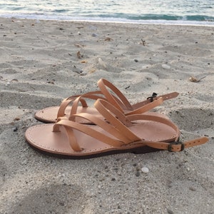 Op de afbeelding: Een paar beige leren sandalen met meerdere dunne bandjes die over de voet lopen, een teenlus en een verstelbaar enkelbandje met een kleine metalen gesp. De sandalen hebben een platte, donkerbruine zool met zichtbare witte stiksels.