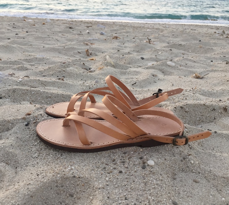 Op de afbeelding: Een paar beige leren sandalen met meerdere dunne bandjes die over de voet lopen, een teenlus en een verstelbaar enkelbandje met een kleine metalen gesp. De sandalen hebben een platte, donkerbruine zool met zichtbare witte stiksels.