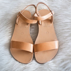 Peut inclure: Une paire de sandales en cuir beige avec une large bride avant et une bride de cheville r&eacute;glable dot&eacute;e d'une boucle en m&eacute;tal argent&eacute;. La semelle int&eacute;rieure porte l'inscription 'REAL LEATHER', 'MADE IN GREECE' et le chiffre 37 dans un cercle.