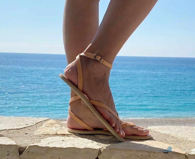 May include: Tan leather sandals with a thin strap across the foot and an ankle strap. The sandals are worn by a person standing on a stone wall with the ocean in the background.