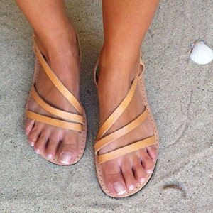 May include: A pair of light tan leather sandals featuring three thin, diagonal straps across the top of the foot and a single heel strap. The sandals have a flat sole with visible stitching along the edges, providing a simple and functional design.