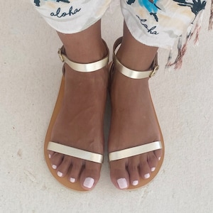 May include: A pair of flat sandals featuring a metallic gold finish, a thin horizontal strap across the forefoot, and an adjustable ankle strap with a small gold-toned metal buckle. The sandals have a simple, open-toe design with a tan-colored sole.