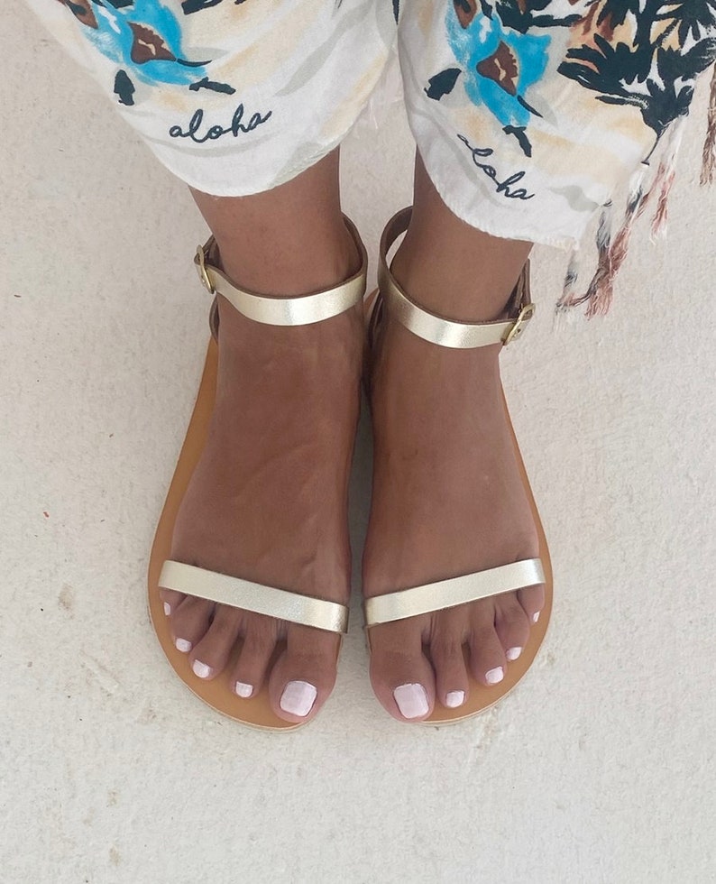 May include: A pair of flat sandals featuring a metallic gold finish, a thin horizontal strap across the forefoot, and an adjustable ankle strap with a small gold-toned metal buckle. The sandals have a simple, open-toe design with a tan-colored sole.