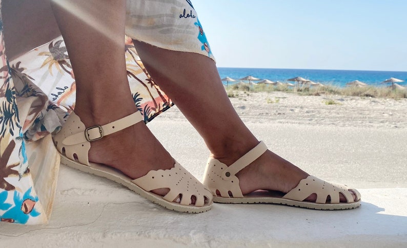 May include: A pair of cream-colored flat sandals featuring a scalloped edge design, cut-out details across the top, and an adjustable ankle strap with a metallic buckle. The sandals have a textured sole and a rounded toe construction.