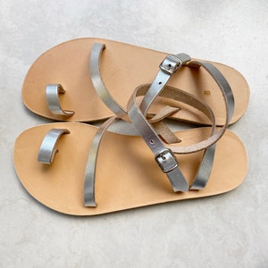May include: A pair of flat sandals featuring light tan leather footbeds and metallic silver leather straps. The design includes a toe loop, thin ankle straps with silver-toned metal buckles, and the number 9 stamped on the heel area of the footbed.