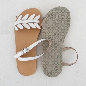 May include: A pair of flat sandals featuring a tan leather footbed and white leather straps. One sandal displays a decorative leaf-shaped cutout design across the top, while the other shows the grey textured rubber sole with a geometric pattern.