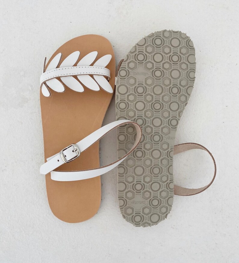 May include: A pair of flat sandals featuring a tan leather footbed and white leather straps. One sandal displays a decorative leaf-shaped cutout design across the top, while the other shows the grey textured rubber sole with a geometric pattern.
