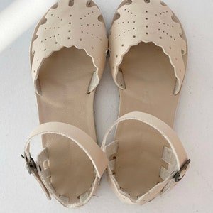 May include: A pair of cream-colored sandals featuring a closed-toe design with decorative scalloped edges and perforated dot patterns. The sandals include an adjustable ankle strap with a small metal buckle, providing a secure fit for the wearer.