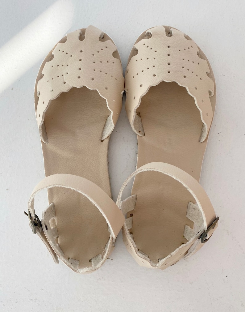May include: A pair of cream-colored sandals featuring a closed-toe design with decorative scalloped edges and perforated dot patterns. The sandals include an adjustable ankle strap with a small metal buckle, providing a secure fit for the wearer.