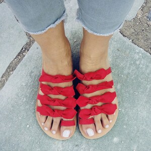 May include: Red leather sandals with multiple bows on each strap. The sandals are worn on a person's feet.