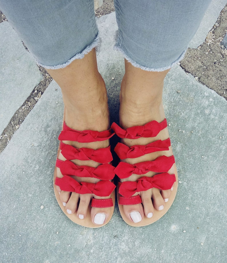 May include: Red leather sandals with multiple bows on each strap. The sandals are worn on a person's feet.