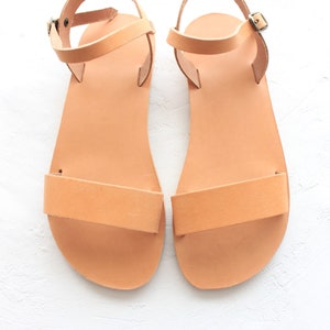May include: A pair of tan leather sandals with ankle straps and buckles. The sandals have a simple design and are made of a soft, supple leather.