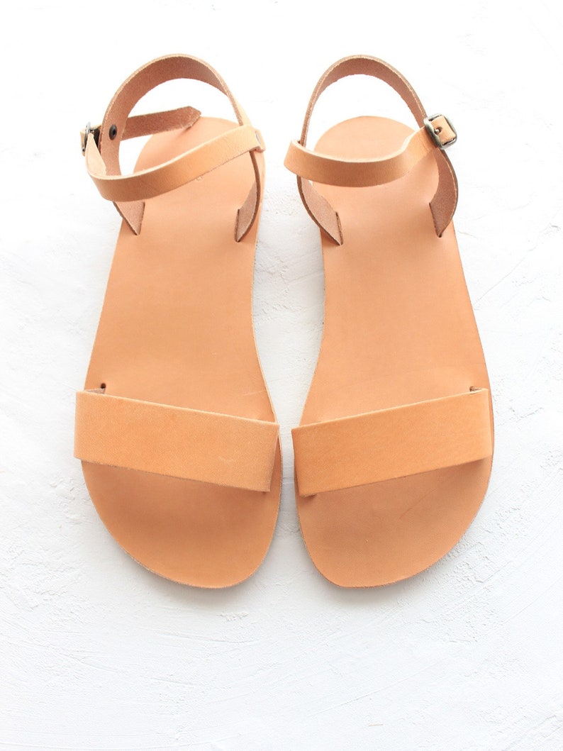 May include: A pair of tan leather sandals with ankle straps and buckles. The sandals have a simple design and are made of a soft, supple leather.