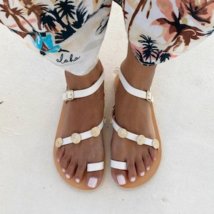May include: White leather sandals featuring a toe loop, an adjustable ankle strap with a gold-tone buckle, and a diagonal strap across the foot adorned with three circular gold-tone metal embellishments with a textured, concentric pattern.