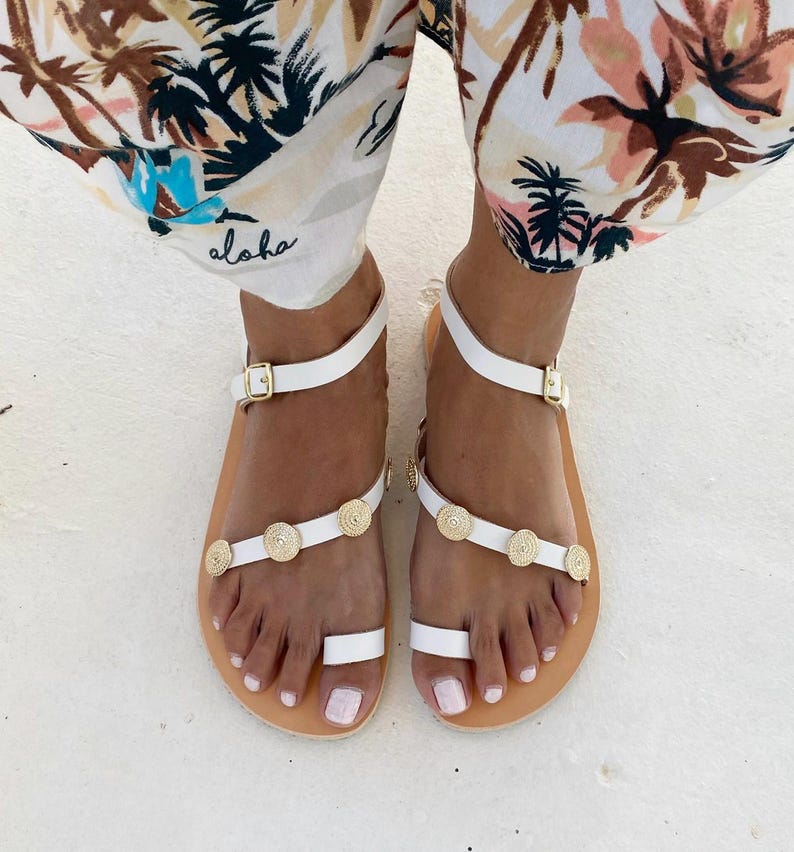 May include: White leather sandals featuring a toe loop, an adjustable ankle strap with a gold-tone buckle, and a diagonal strap across the foot adorned with three circular gold-tone metal embellishments with a textured, concentric pattern.