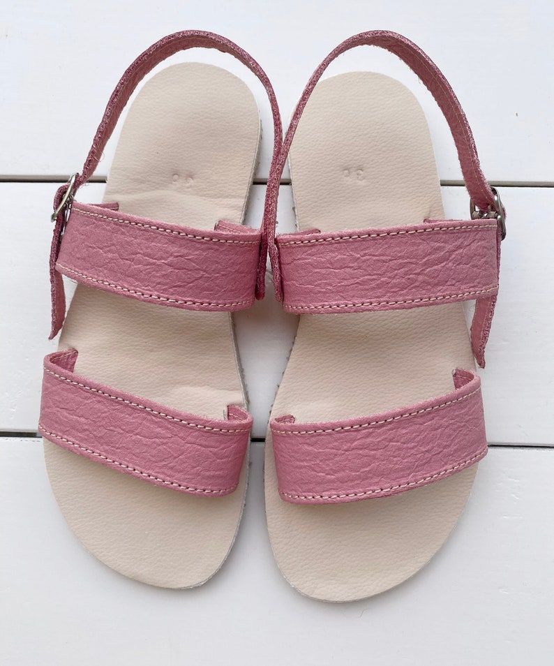 May include: A pair of dusty rose sandals featuring two wide textured straps across the top and an adjustable ankle strap with a silver-toned metal buckle. The footbed is a light beige color, and the number 36 is embossed on the heel area.
