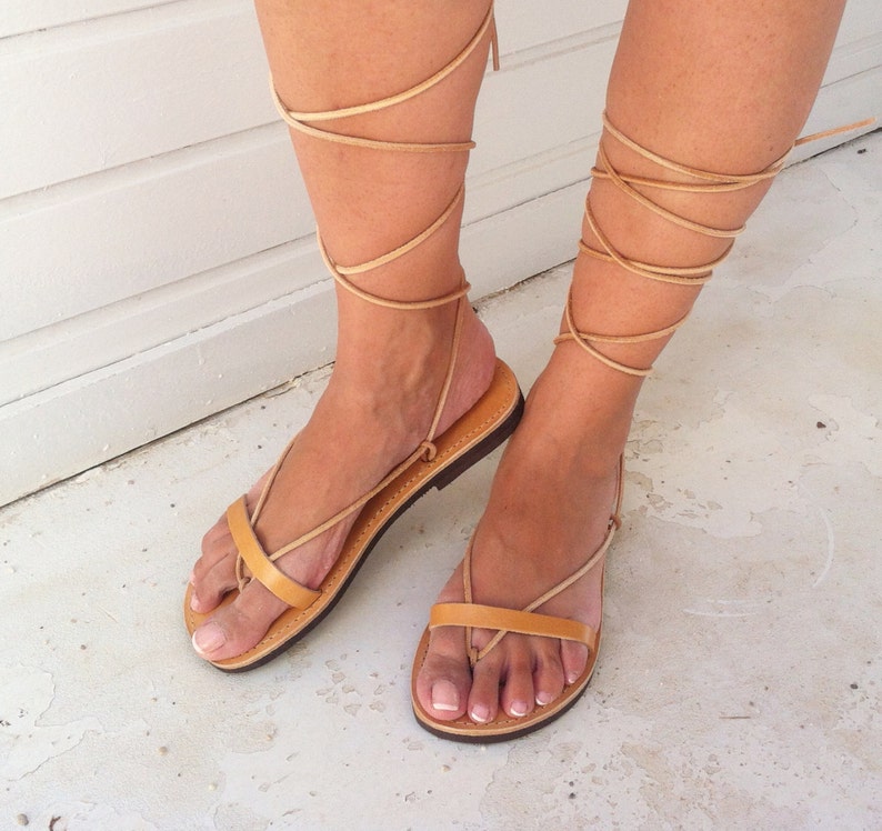 May include: Tan leather sandals with laces that wrap up the legs. The sandals have a flat sole and a simple design.