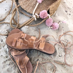 Op de afbeelding: Een paar beige leren vetersandalen met platte zolen en een ronde tas van gevlochten stro, versierd met een snoer van lichte houten kralen, drie oudroze garen pompons en een grote bijpassende kwast bevestigd aan het bovenste handvat.