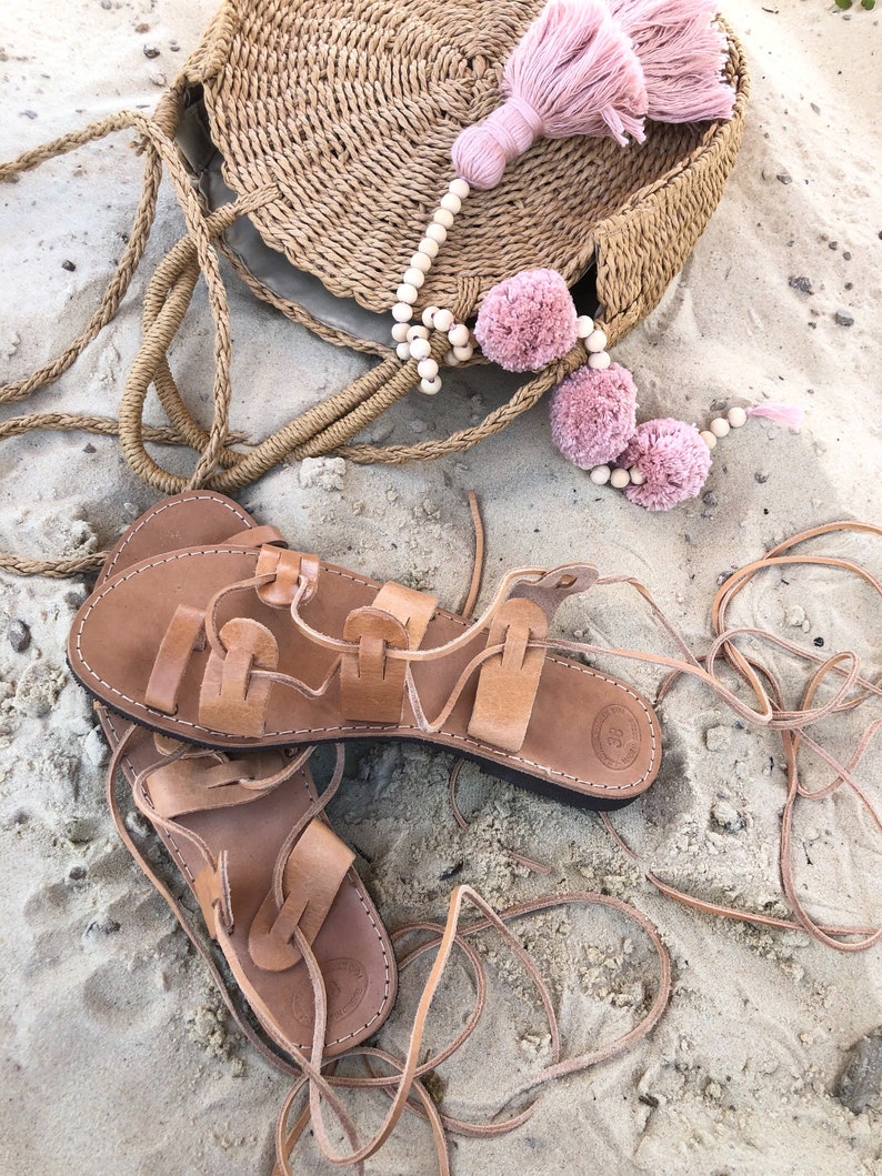 Op de afbeelding: Een paar beige leren vetersandalen met platte zolen en een ronde tas van gevlochten stro, versierd met een snoer van lichte houten kralen, drie oudroze garen pompons en een grote bijpassende kwast bevestigd aan het bovenste handvat.