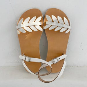 May include: A pair of flat sandals featuring a tan leather footbed and a white patent leather strap across the top adorned with a series of white leaf-shaped cutouts. The sandals include a thin white ankle strap with a silver-toned metal buckle.