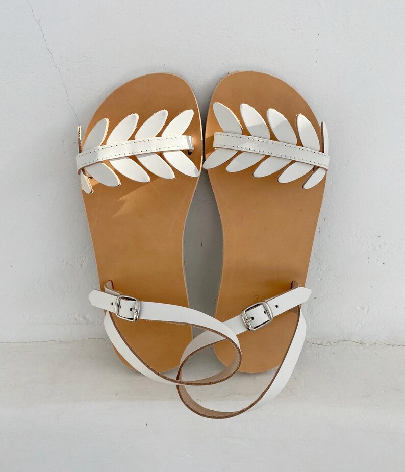 May include: A pair of flat sandals featuring a tan leather footbed and a white patent leather strap across the top adorned with a series of white leaf-shaped cutouts. The sandals include a thin white ankle strap with a silver-toned metal buckle.