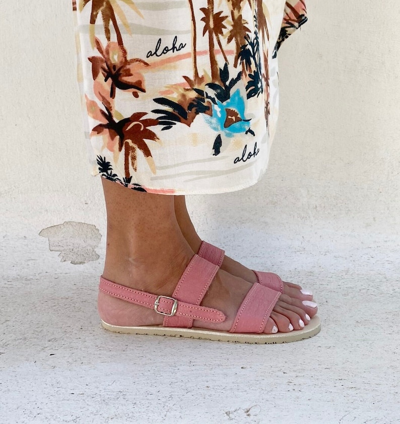 May include: A pair of pink flat sandals with two horizontal straps and a thin ankle strap featuring a silver-toned metal buckle. The sandals have a light-colored sole. Above the sandals, a cream-colored fabric features a brown and blue palm tree print and the text aloha.