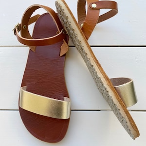 Peut inclure: Une paire de sandales plates avec une bride avant en cuir dor&eacute; m&eacute;tallis&eacute; et une bride de cheville marron dot&eacute;e d'une boucle en m&eacute;tal dor&eacute;. La semelle int&eacute;rieure est marron fonc&eacute; et la semelle ext&eacute;rieure pr&eacute;sente un motif textur&eacute;.