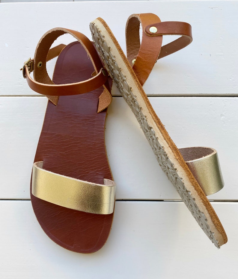 Peut inclure: Une paire de sandales plates avec une bride avant en cuir dor&eacute; m&eacute;tallis&eacute; et une bride de cheville marron dot&eacute;e d'une boucle en m&eacute;tal dor&eacute;. La semelle int&eacute;rieure est marron fonc&eacute; et la semelle ext&eacute;rieure pr&eacute;sente un motif textur&eacute;.