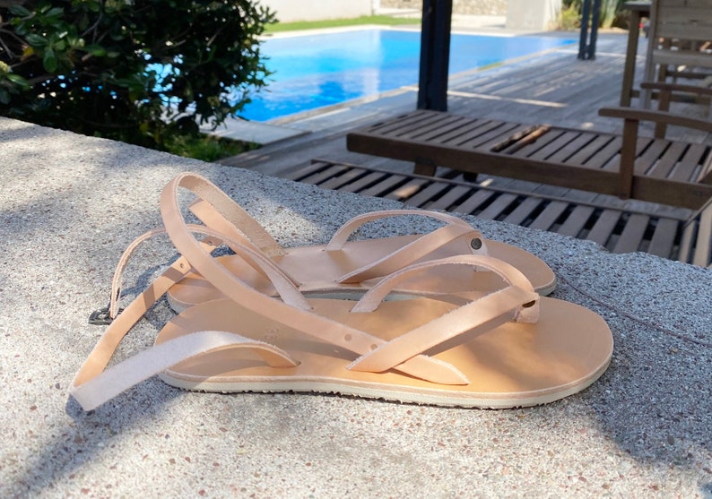 May include: Two pairs of tan leather sandals with adjustable straps. The sandals are lying on a concrete surface near a swimming pool.