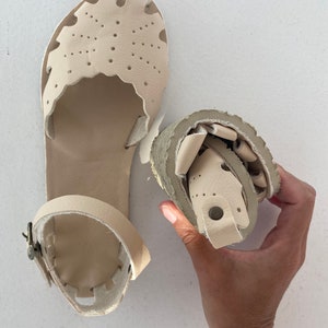 May include: A pair of beige leather sandals featuring a closed-toe design with decorative cutouts and perforations. One sandal is laid flat, showing the ankle strap with a metal buckle, while the other is folded to demonstrate flexibility.