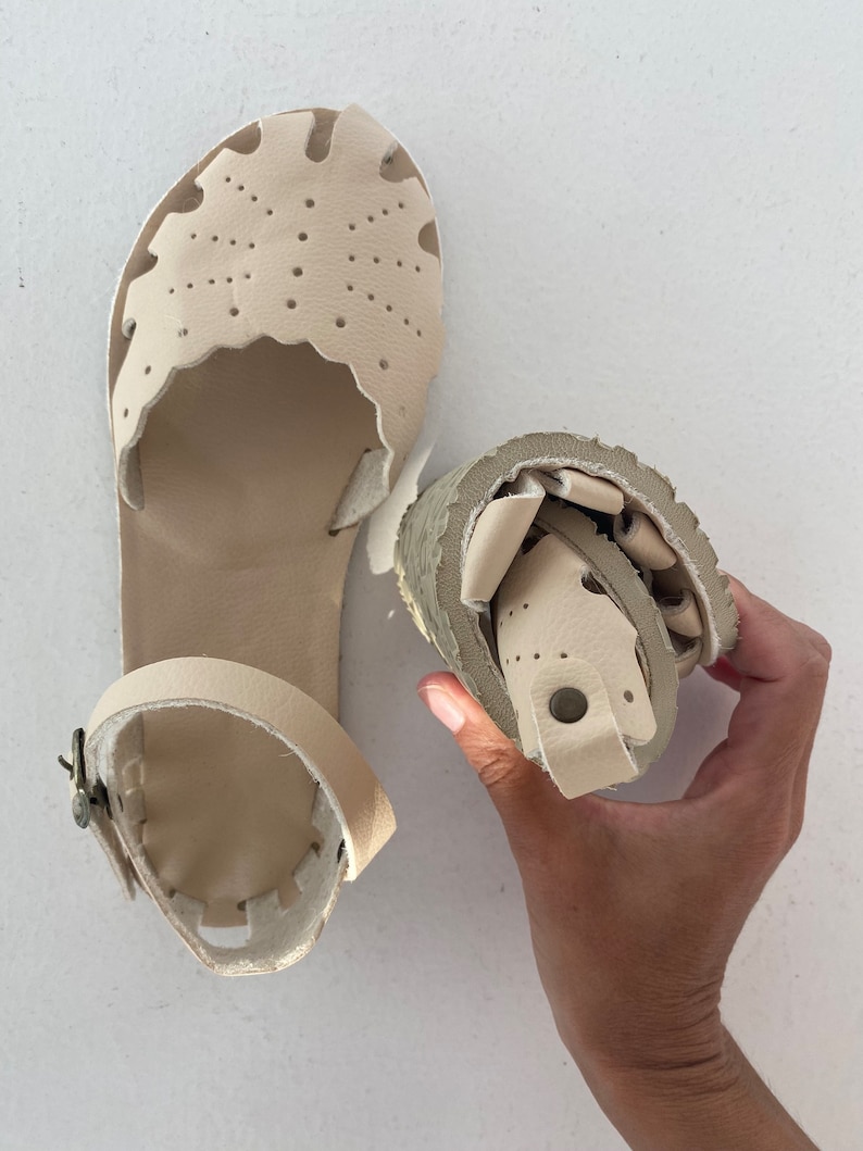 May include: A pair of beige leather sandals featuring a closed-toe design with decorative cutouts and perforations. One sandal is laid flat, showing the ankle strap with a metal buckle, while the other is folded to demonstrate flexibility.