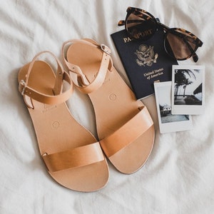 May include: Tan leather sandals with a buckle closure, a passport with the text "PASSPORT" and "United States of America", sunglasses, and two instant photos of a tropical beach scene.