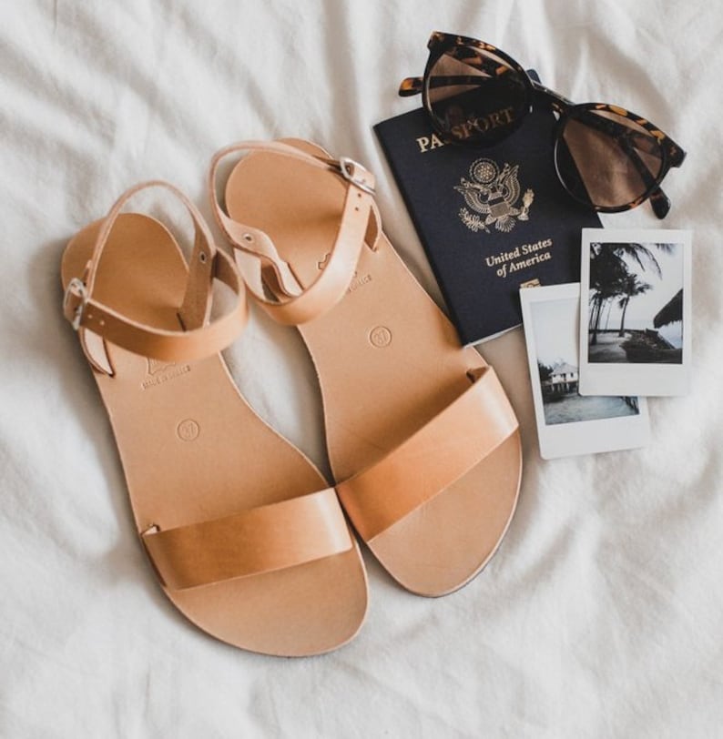 May include: Tan leather sandals with a buckle closure, a passport with the text "PASSPORT" and "United States of America", sunglasses, and two instant photos of a tropical beach scene.