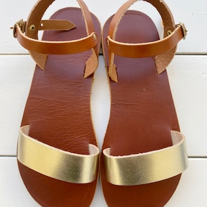 Peut inclure: Une paire de sandales plates dot&eacute;es d'une semelle int&eacute;rieure en cuir marron et d'une bride dor&eacute;e m&eacute;tallique sur l'avant-pied. Chaque sandale comprend une fine bride de cheville en cuir marron avec une petite boucle en m&eacute;tal dor&eacute;.