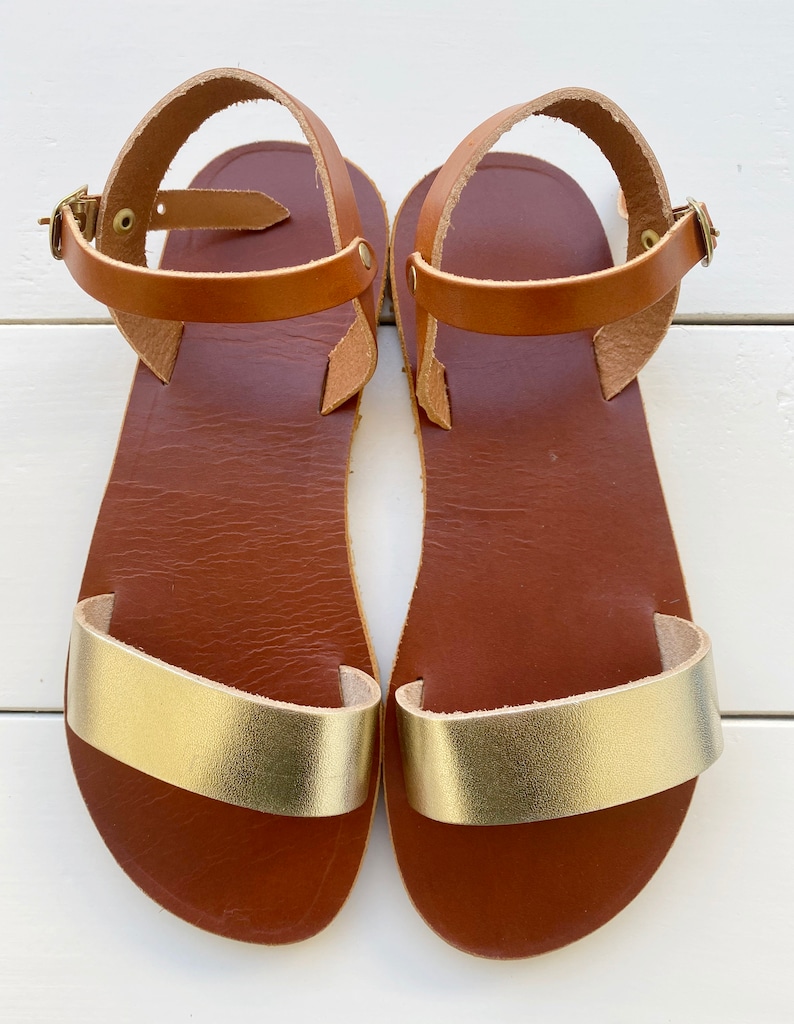 Peut inclure: Une paire de sandales plates dot&eacute;es d'une semelle int&eacute;rieure en cuir marron et d'une bride dor&eacute;e m&eacute;tallique sur l'avant-pied. Chaque sandale comprend une fine bride de cheville en cuir marron avec une petite boucle en m&eacute;tal dor&eacute;.