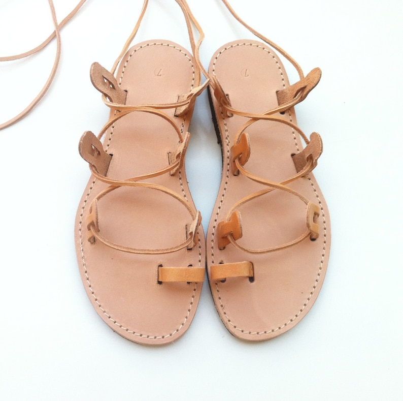 Op de afbeelding: Een paar beige leren sandalen met een teenlus, meerdere zijbandjes en lange veters om rond de enkel te wikkelen. Het voetbed is gestempeld met het cijfer 7 en de randen zijn afgewerkt met zichtbare witte stiksels langs de zool.